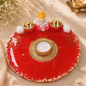 Round red resin pooja thali with a jagged gold-flecked edge, featuring a  Ganesha figurine, two small gold diyas, white rose accents, and gold leaf decorations, with a large ornate gold tealight holder centered on the tray.