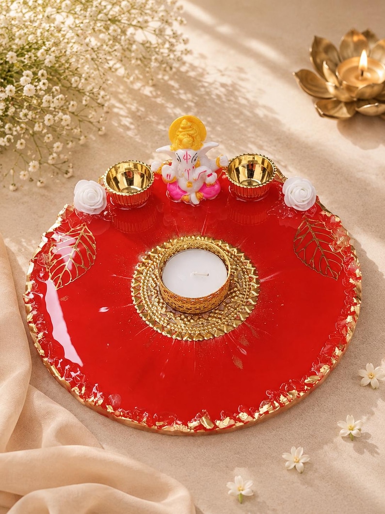 Round red resin pooja thali with a jagged gold-flecked edge, featuring a  Ganesha figurine, two small gold diyas, white rose accents, and gold leaf decorations, with a large ornate gold tealight holder centered on the tray.