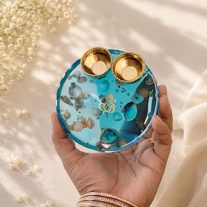 Round turquoise blue resin pooja thali embedded with seashells and a gold Ganesh emblem in the center, topped with two small gold diyas.