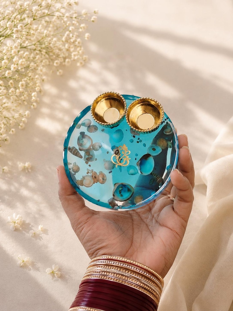 Round turquoise blue resin pooja thali embedded with seashells and a gold Ganesh emblem in the center, topped with two small gold diyas.