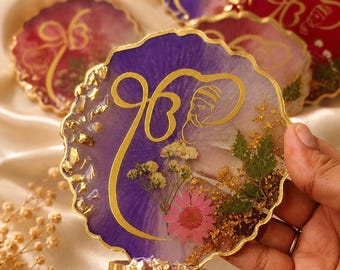 May include: A round, scalloped-edge resin coaster featuring a purple and white gradient background, gold leaf accents, and embedded dried flowers including a pink daisy. A gold-coloured Ik Onkar symbol is printed on the surface.