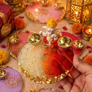May include: A circular resin tray featuring a central white and orange Ganesha figurine, two gold-toned metal diya cups, and a gold swastika symbol. The tray is filled with white rice on one side and red pigment on the other, with gold leaf flakes along the edge.