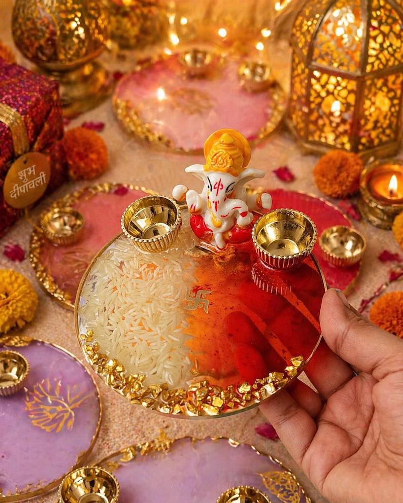 May include: A circular resin tray featuring a central white and orange Ganesha figurine, two gold-toned metal diya cups, and a gold swastika symbol. The tray is filled with white rice on one side and red pigment on the other, with gold leaf flakes along the edge.
