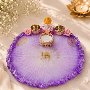 Round purple and white marbled resin pooja thali with a jagged crystal-flecked edge, featuring a white and gold Ganesha figurine, a white tealight candle in a gold holder, two small gold diyas, pink rose accents, and gold leaf decorations, with a gold swastik symbol at the center of the tray.