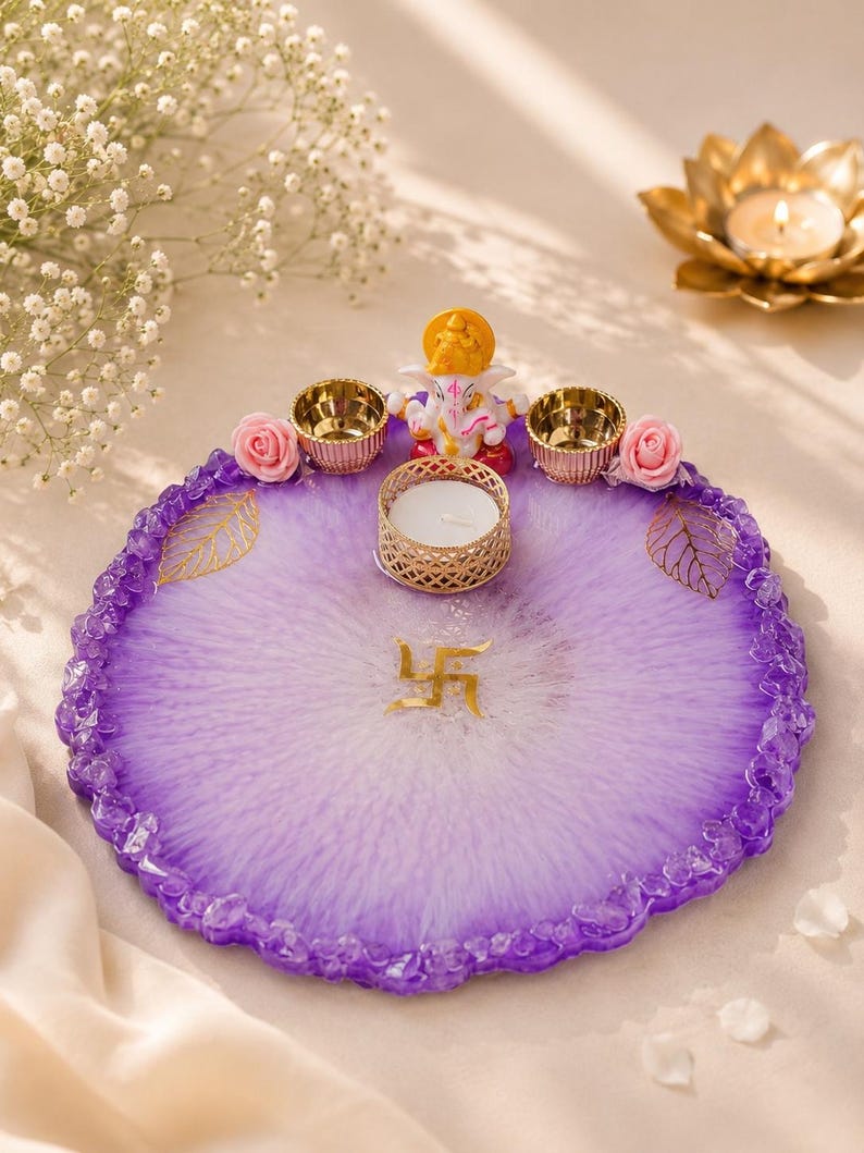 Round purple and white marbled resin pooja thali with a jagged crystal-flecked edge, featuring a white and gold Ganesha figurine, a white tealight candle in a gold holder, two small gold diyas, pink rose accents, and gold leaf decorations, with a gold swastik symbol at the center of the tray.