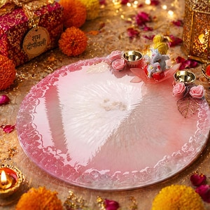 May include: A circular pink and white resin tray featuring a textured, translucent finish. The tray is decorated with a small figurine of Ganesha, two metallic gold-toned diya candle holders, pink artificial roses, and gold-toned leaf accents.
