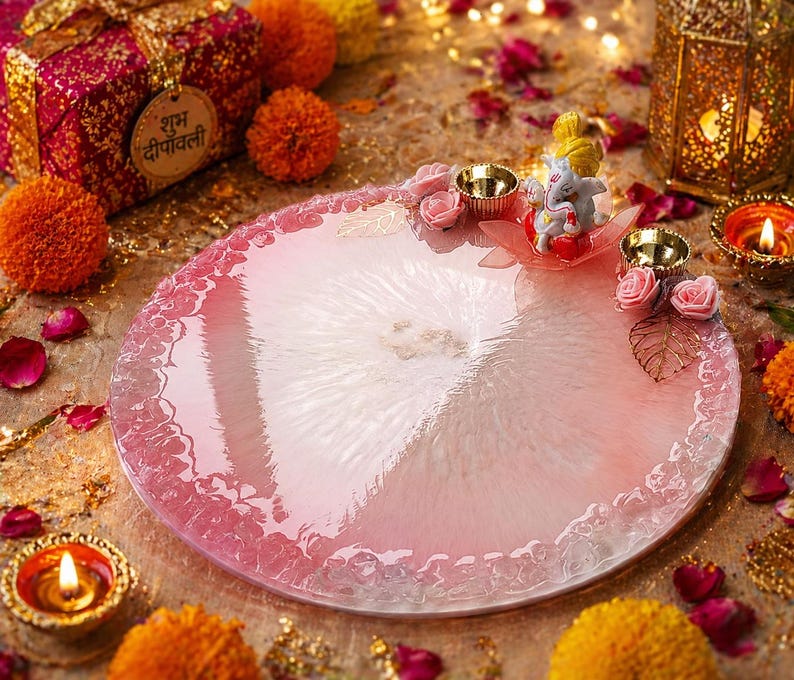 May include: A circular pink and white resin tray featuring a textured, translucent finish. The tray is decorated with a small figurine of Ganesha, two metallic gold-toned diya candle holders, pink artificial roses, and gold-toned leaf accents.