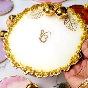 Hand holding a white resin pooja thali with a jagged gold-flecked edge, gold leaf accents, small gold diyas, and a gold Ik Omkar emblem etched in the center.