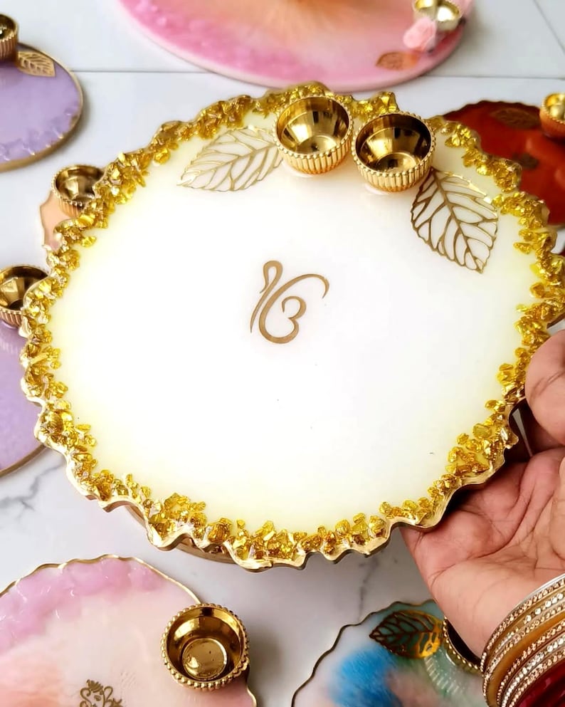 Hand holding a white resin pooja thali with a jagged gold-flecked edge, gold leaf accents, small gold diyas, and a gold Ik Omkar emblem etched in the center.