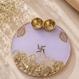 Round lavender resin pooja thali with a gold-flecked design along the bottom edge, featuring two small gold diyas, gold leaf decorations, and a gold swastik symbol at the center.