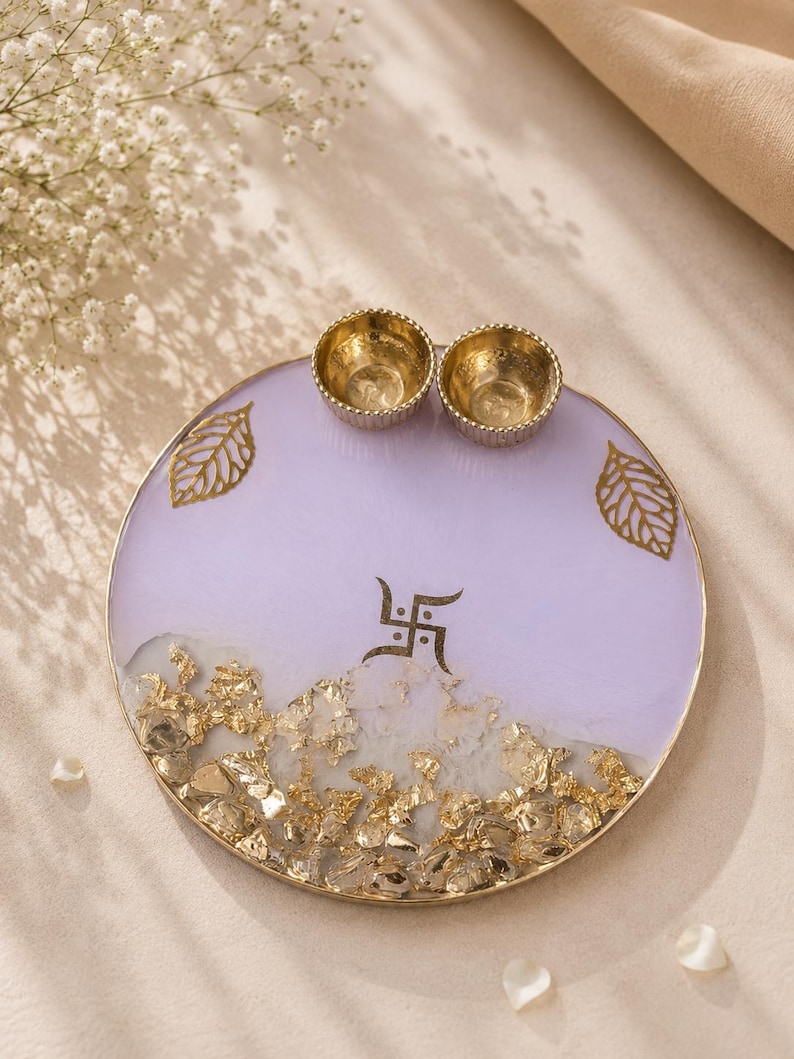 Round lavender resin pooja thali with a gold-flecked design along the bottom edge, featuring two small gold diyas, gold leaf decorations, and a gold swastik symbol at the center.