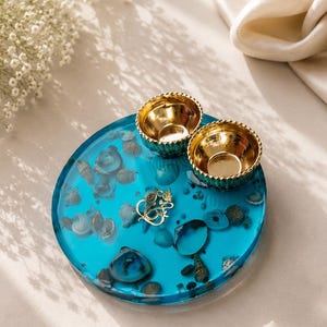 Round turquoise blue resin pooja thali embedded with seashells and a gold Ganesh emblem in the center, topped with two small gold diyas.