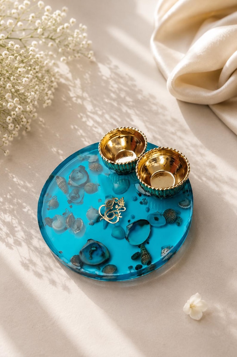 Round turquoise blue resin pooja thali embedded with seashells and a gold Ganesh emblem in the center, topped with two small gold diyas.