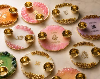 May include: A collection of circular resin decorative plates, each featuring two small gold-toned metal bowls. The plates display various colours, including pink, white, green, and orange, with gold-leaf accents and printed symbols or names like Arpit, Shruti, Ayush, and Surbhi.