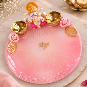 May include: A circular pink and white resin tray featuring a textured, translucent finish. The tray is decorated with a small figurine of Ganesha, two metallic gold-toned diya candle holders, pink artificial roses, and gold-toned leaf accents.