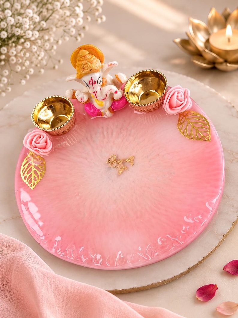 May include: A circular pink and white resin tray featuring a textured, translucent finish. The tray is decorated with a small figurine of Ganesha, two metallic gold-toned diya candle holders, pink artificial roses, and gold-toned leaf accents.