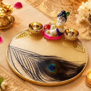 Round gold-rimmed pooja thali with an embedded peacock feather design, decorated with two small gold diyas and a Baby Krishna figurine seated at the top of the tray.