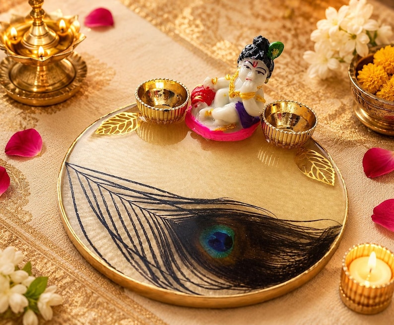 Round gold-rimmed pooja thali with an embedded peacock feather design, decorated with two small gold diyas and a Baby Krishna figurine seated at the top of the tray.