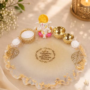 Round cream and gold resin pooja thali with a wavy gold-flecked edge, featuring a Ganesha figurine, a white tealight candle in a gold holder, two small gold diyas, white rose accents, and gold leaf decorations, with a Gayatri mantra inscribed in a decorative circular border at the center.