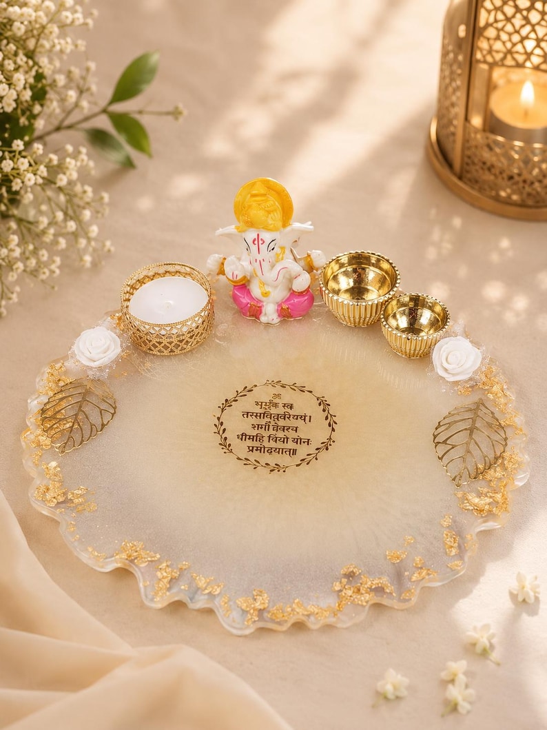 Round cream and gold resin pooja thali with a wavy gold-flecked edge, featuring a Ganesha figurine, a white tealight candle in a gold holder, two small gold diyas, white rose accents, and gold leaf decorations, with a Gayatri mantra inscribed in a decorative circular border at the center.