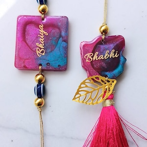 May include: Two decorative hanging ornaments featuring pink and blue resin pieces. One is a square with the text Bhaiya, the other is an irregular shape with the text Bhabhi. Both include gold-toned beads, a gold leaf charm, and a pink tassel.