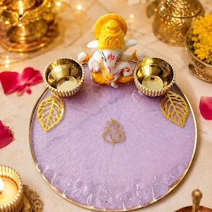 A circular lavender resin tray with a gold-toned rim, featuring a smallGanesha figurine at the top, two gold-toned metal bowls, two gold leaf-shaped accents, and a central gold Ganesha symbol emblem.