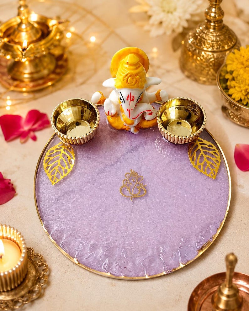 A circular lavender resin tray with a gold-toned rim, featuring a smallGanesha figurine at the top, two gold-toned metal bowls, two gold leaf-shaped accents, and a central gold Ganesha symbol emblem.