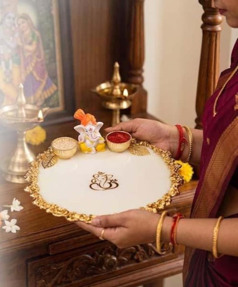 A white resin pooja thali with gold-toned decorative edges and a central gold Ganesha emblem. The tray features a small, colourful Ganesha figurine flanked by two gold-toned bowls.