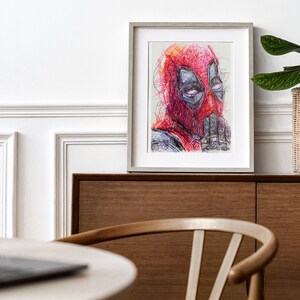 May include: Framed artwork featuring a colorful, abstract portrait of Deadpool. The artwork is predominantly red, with details in black, gray, and purple. The framed print is displayed on a wooden cabinet, with a white wall in the background.