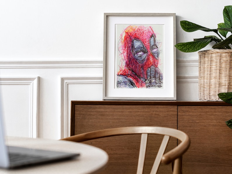 May include: Framed artwork featuring a colorful, abstract portrait of Deadpool. The artwork is predominantly red, with details in black, gray, and purple. The framed print is displayed on a wooden cabinet, with a white wall in the background.