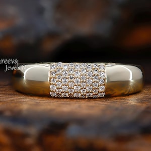 May include: A gold ring featuring a central rectangular section encrusted with small, clear gemstones. The ring has a polished finish and is presented on a wooden surface. The ring is a piece of jewellery.