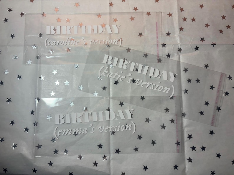 May include: Three clear plastic bags with white text that reads "BIRTHDAY (caroline's version)", "BIRTHDAY (katie's version)", and "BIRTHDAY (emma's version)". The bags are on a white background with silver glitter stars.