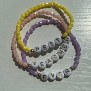 May include: Three beaded bracelets in shades of yellow, pink, and purple. The bracelets are strung with small, faceted beads. One bracelet spells out "BEST TEACHER EVER" with white letter beads and gold accents. A thoughtful gift.