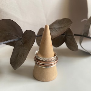 Puede incluir: Un conjunto de tres anillos de metal martillados, uno plateado y dos de cobre, apilados en un soporte de anillo de madera.