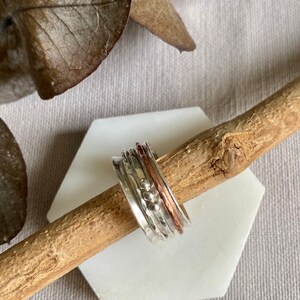 Puede incluir: Un anillo giratorio de plata con detalles de cobre y cuentas. El anillo se muestra sobre una superficie hexagonal blanca, apoyado en una ramita de madera, con hojas secas en el fondo. El diseño del anillo incluye múltiples bandas giratorias.