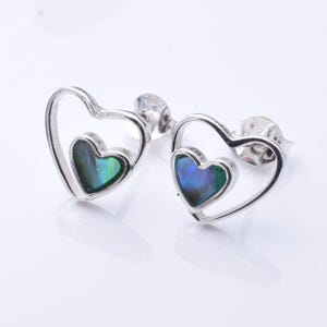 May include: A pair of heart-shaped stud earrings. Each earring has a silver heart outline, with a smaller heart-shaped inlay of iridescent blue and green material. The earrings are displayed on a white surface.