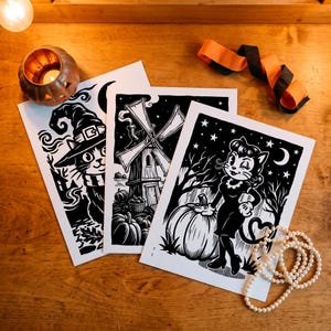 May include: Three rectangular white paper prints featuring black ink illustrations. One print shows a cat wearing a witch hat, another depicts a windmill with pumpkins, and the third displays a cat character standing next to a large pumpkin.