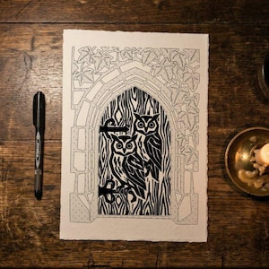 May include: A black and white linocut print on textured paper featuring two owls perched within a gothic archway surrounded by ivy leaves. The artwork is displayed alongside a black ink pen and a small brass bowl containing a candle.