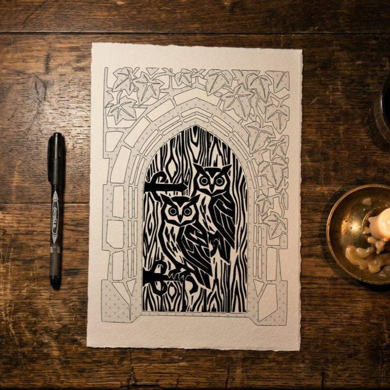 May include: A black and white linocut print on textured paper featuring two owls perched within a gothic archway surrounded by ivy leaves. The artwork is displayed alongside a black ink pen and a small brass bowl containing a candle.