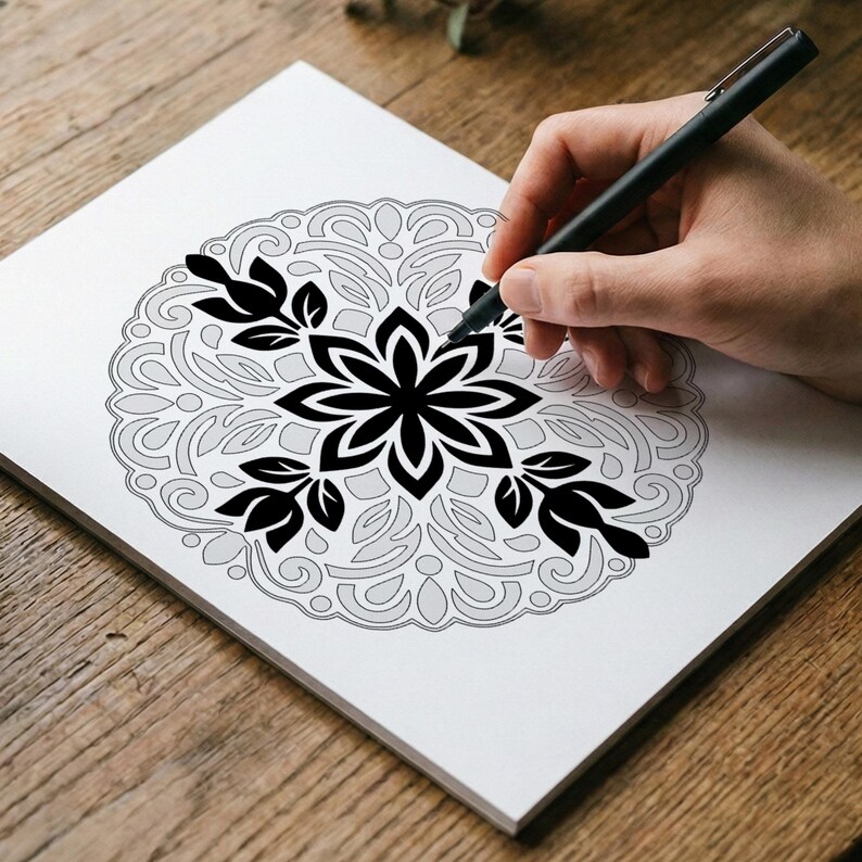 May include: A black pen drawing a detailed floral mandala design on white paper. The mandala features a central flower shape with surrounding leaf and petal patterns. The design is partially shaded in gray.