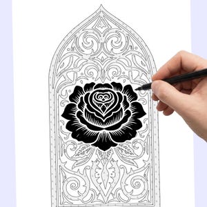 May include: A black rose design within a decorative arched frame, possibly for a coloring book or craft project. The image is in black and white, with a hand holding a pen, suggesting the design is being drawn or colored.