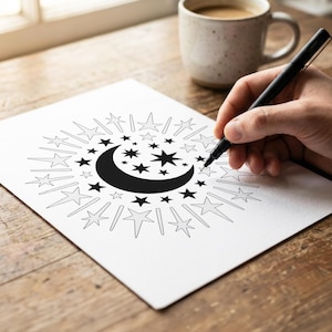 May include: A black and white celestial design featuring a crescent moon and stars, surrounded by starbursts, is being drawn on white paper with a black pen. A mug of coffee sits nearby on a wooden surface.