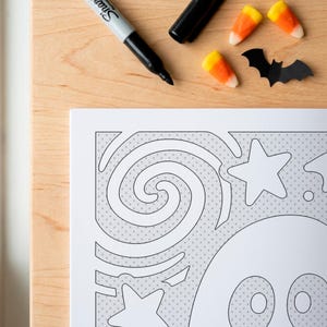May include: A black Sharpie marker, two orange and yellow candy corn pieces, and a black paper bat cutout arranged near a white coloring page featuring a ghost, stars, and swirling lines with a light gray cross-hatched background pattern.