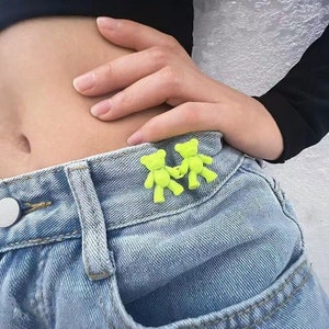 May include: Two neon green teddy bear shaped belt clips attached to a pair of light blue denim jeans.