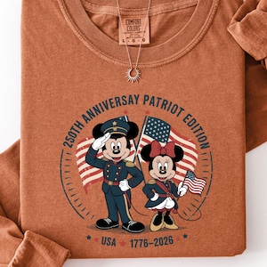 a brown t-shirt with a graphic design depicting two cartoon characters, Mickey Mouse and Minnie Mouse, dressed in military uniforms and holding American flags. The text on the t-shirt reads "50th Anniversary Patriotic Edition USA 1776-2025".