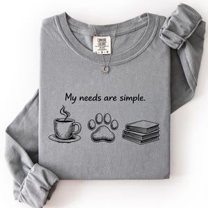May include: A grey long-sleeve shirt with the text "My needs are simple." The graphic includes a coffee cup, a paw print, and two books. The shirt is made of a soft material.