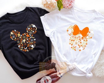 May include: Black and white t-shirts featuring a Halloween-themed Mickey Mouse design. The Mickey Mouse silhouette is filled with orange, white, and yellow Halloween icons. The white shirt has an orange bow.