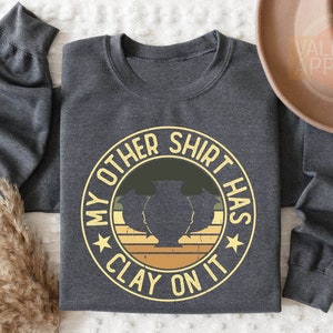 May include: Dark heather gray sweatshirt with a circular graphic that reads "MY OTHER SHIRT HAS CLAY ON IT." The design features a pottery vase silhouette in green and yellow tones, with a retro sunset design. The text is in a yellow and white color.