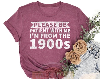 May include: Heather maroon t-shirt with the text "Please be patient with me I'm from the 1900s" in white. The shirt is tied in a knot at the bottom and is paired with light blue denim shorts.
