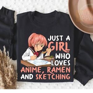 May include: Black t-shirt with a graphic of a girl with pink hair eating ramen and drawing. The text on the shirt reads "Just a girl who loves anime, ramen and sketching."